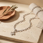 A silver chain necklace with a small textured animal charm, styled on a beige stone slab with a terracotta plate, dried leaves, and a rolled cream fabric.